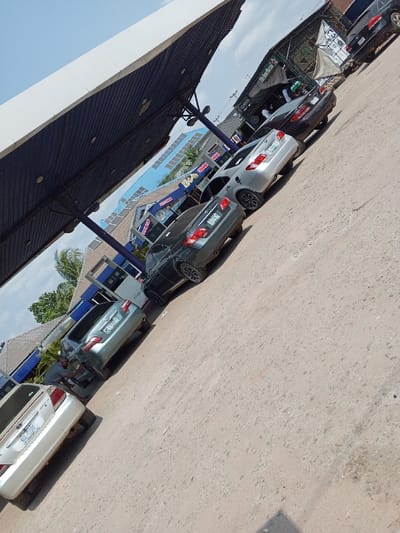 The image depicts an outdoor service station scene in Naze, Nigeria, under a bright sky with scattered clouds. The ground is unpaved, dusty, and light brown. A blue and white corrugated metal canopy, supported by blue pillars, covers the left side of the frame. Five sedans are arranged in a queue, facing away from the viewer. From left to right, these vehicles are: a white sedan (license plate KGC 338FF), a light grey sedan (license plate LAG 244EX), a dark grey sedan (license plate LAG 392ES), a silver sedan (license plate LAG 546CA), and a partially visible dark sedan on the far right. A male individual, wearing a grey t-shirt and dark trousers, is kneeling or bending by the rear passenger side of the light grey sedan (LAG 244EX), appearing to perform a task. A white fuel dispenser, marked with "7739", is positioned between the light grey and dark grey sedans. In the background, a building with a light blue roof fitted with multiple solar panels and signage including "SHARE" is visible. Adjacent to it, a green-roofed structure displaying a "Heineken" banner shelters several indistinct individuals. Palm trees are visible in the distant background.
