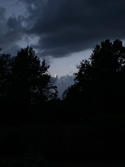 The image depicts a dark, silhouetted landscape with a sliver of a crescent moon visible in the night sky. The sky is filled with heavy, dark clouds. The foreground is completely black, suggesting it is nighttime. Trees with dense foliage are silhouetted against the sky on either side of the frame. The scene appears to be a natural outdoor setting, possibly a forest or park, at dusk or night. There is no indication of human presence or activity in the image. The location is stated to be the city of Courpière, France.