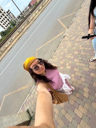A first-person perspective, selfie-style image captures a young woman wearing a yellow headscarf, oversized sunglasses, and a pink top with a white skirt. She is holding a brown shoulder bag. The woman is standing on a paved walkway with multi-colored bricks, adjacent to a paved road with yellow markings. In the background, a set of train tracks runs parallel to the road, with a partially constructed building to the left and some houses to the right. Another person, partially visible on the right side of the frame, is wearing a blue outfit and black flip-flops, holding a black object. The scene appears to be an urban setting in Derbent, Russia, with a clear sky overhead.