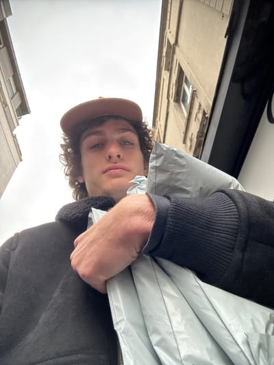 A low-angle, upward-facing image captures a young male individual in an urban environment in Joinville-le-Pont, France, under an overcast sky. The subject, centrally framed, has curly dark brown hair, blue eyes, and a neutral expression, gazing directly at the viewer. He wears a brown baseball cap and a dark-colored jacket with a black sherpa-style collar and ribbed cuffs. His left hand, positioned in the lower right foreground, firmly grips a crinkled, light grey, possibly plastic or synthetic, package or bag. The background shows two multi-story buildings flanking the open sky: a light-colored building with multiple windows on the left and a stone-textured building with similar windows and a dark vertical feature on the lower right on the right.