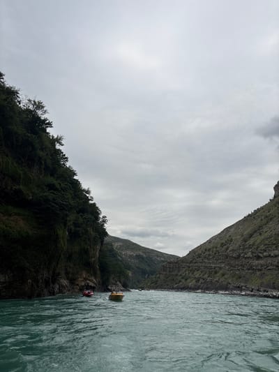 The image captures a wide, turquoise river flowing through a canyon. Steep, densely vegetated cliffs rise on either side of the water. The sky is overcast with gray clouds. Two small boats are visible on the river. One is a red raft with people, and the other is a yellow motorboat. The scene suggests outdoor recreation or transport on the river within a mountainous, natural landscape, possibly near Chirkey, Russia, given the context provided.