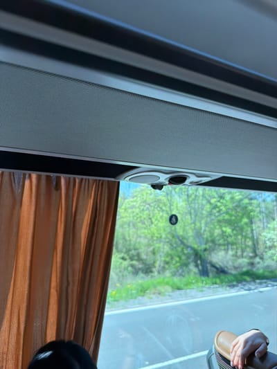 The image shows an interior view, likely taken from a bus or train in Prilep, Bulgaria, based on the scenery visible through the window. The window is positioned to show a blurred view of trees and a road outside. A section of the vehicle's interior is visible: a gray wall or panel and a brown curtain. In the upper right corner, there is a glimpse of a person's hand and part of a shoe or sandal.