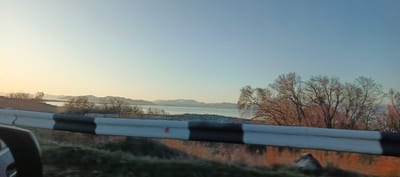 The image presents a tranquil, expansive landscape viewed from a moving vehicle, likely during sunrise or sunset. In the foreground, the edge of a car's side mirror is visible on the left, along with a black and white striped guardrail running horizontally across the lower part of the frame, indicating motion blur.

Beyond the guardrail, a natural scene unfolds. The midground is dominated by bare deciduous trees, their branches appearing dry and stark, suggesting a late autumn or winter season. Behind the trees, a wide body of water, likely a lake or large river, stretches towards the horizon.

In the background, a majestic range of mountains rises. Several peaks, particularly towards the right, are visibly capped with snow. The sky above is clear and vast, exhibiting a gentle gradient of color: a soft, warm yellow and orange on the left transitions smoothly into a pale blue on the right, characteristic of either early morning or late afternoon light. A faint, thin contrail from an aircraft is visible high in the upper right sky.

The scene conveys a sense of peaceful natural beauty, common in mountainous regions. The location context of Berdkunk, Armenia, fits this description of mountains and a body of water. There is no visible text in the image.
