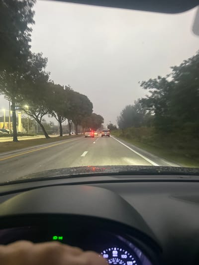 The image depicts a first-person perspective from inside a vehicle traveling eastbound on NW 41st Street in Doral, Florida, United States of America, in the vicinity of NW 97th Avenue. The road surface is wet, indicating recent precipitation, under a uniformly overcast, gray sky.

In the foreground, the lower portion of a vehicle's dashboard, a segment of the steering wheel, and a human left hand are visible. A green indicator light, signifying activated headlights, is illuminated on the dashboard. A speedometer/tachometer cluster is partially discernible.

The vehicle is on a multi-lane asphalt road with dashed white lane markings separating the lanes of travel and a solid white line on the right shoulder. Several vehicles are ahead in the same lane, displaying illuminated red taillights. The headlights of the subject vehicle are visible illuminating the wet road surface.

To the left of the road, a landscaped median separates opposing traffic lanes or a commercial roadside. Visible commercial establishments include a building with "Dunkin'" signage and an adjacent "Bank" building, identifiable as a Chase Bank. Multiple trees, including palm trees, line the left side of the road. The right side of the road features a dense line of trees and foliage.