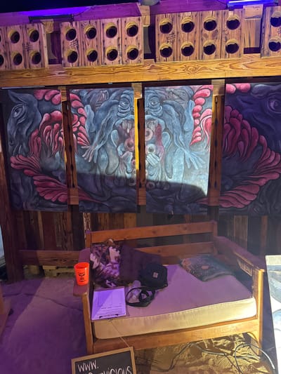 The image captures an interior scene in what appears to be a dimly lit, rustic or bohemian-style space. Located within Joshua Tree, United States, the image is of a quirky gallery and lounge with distinct and unconventional features.

On the left, there's a tall structure made of wooden crates with circular holes, possibly repurposed from wine bottles or similar containers. Adjacent to it, a series of artistic canvases or paintings are stacked vertically against a wooden support. The artwork has abstract or surrealist motifs, with shades of red and blue.

To the right of the canvases is a makeshift lounge area, built up of wooden pallets or planks. A thin mattress is perched on top, with a few pillows and what seems to be a black bag. A clipboard and other personal items are on this elevated surface, and an orange cup is on a shelf nearby. The back of the structure is next to a rough wall and cables/wires. The lighting is predominantly purple/blue, giving the space a unique and artistic vibe.