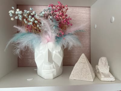 The image shows a shelf in a room. On the shelf are a white geometric vase shaped like a face, filled with dried flowers and feathers in pink, blue, and white. To the right of the vase are two small decorative statues, one a pyramid and the other a sphinx, both appearing to be made of stone with carved details. The background is a wall with a textured pink pattern. The shelf itself is white. There are two white circular adhesive hooks on the right wall of the shelf. A small white circular object, possibly a button or a sticker, is on the shelf in front of the vase. The lighting suggests an indoor environment. There are no people visible in the image. There is no information to connect this scene to Tskhinvali, South Ossetia.