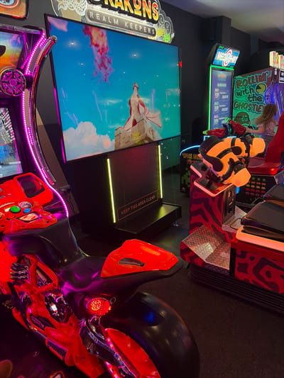 The photo shows an arcade with various games. “Drakons: Realm Keepers” is a game with a large screen displaying a character amidst clouds. Nearby is a motorcycle-shaped game with vibrant red and black accents and a “Keep This Area Clear” sign. In the background, "Rollin' with the Ghosties" is visible with game rankings displayed. A person is near another game, possibly playing. Neon lights illuminate the arcade atmosphere.