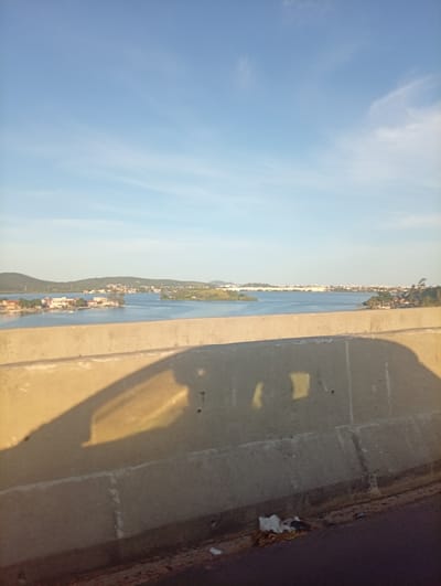 The image presents a panoramic view of a wide body of water, likely Lagoa de Araruama, from an elevated perspective, possibly a bridge or overpass, in São Pedro da Aldeia, Brazil. The foreground is dominated by a sturdy, light-colored concrete barrier that spans the lower half of the frame. A large, elongated shadow of a vehicle, presumably the one from which the photo was taken, is cast across this barrier, stretching from the left side towards the center-right. This shadow, along with the warm light, indicates that the photograph was taken during either the early morning or late afternoon, often referred to as the golden hour.

Beyond the concrete barrier, a vast expanse of calm, blue water fills the midground. Several landmasses, appearing as small islands or peninsulas, emerge from the water. These land areas are visibly populated with numerous houses and buildings, indicating a settled community, interspersed with green trees and vegetation. In the background, rolling hills or mountains covered in greenery can be seen, with additional urban development lining the distant horizon.

The sky is a clear, bright blue, featuring only a few faint, wispy clouds, particularly towards the right side of the frame. The overall weather appears sunny and pleasant. The very bottom edge of the image reveals a dark road surface with some discarded litter beside the concrete barrier. No people or visible text are present in the image. The scene conveys a sense of peaceful expanse and coastal urban life.