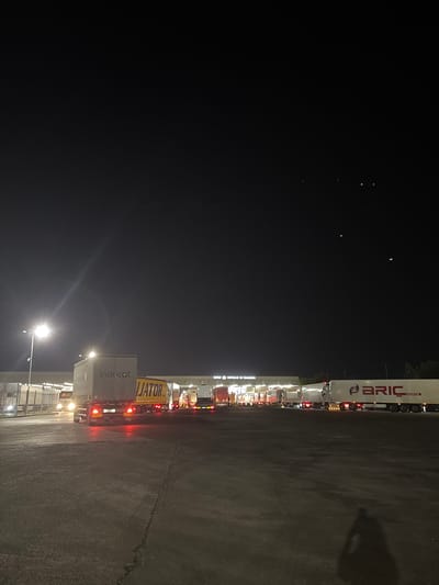 This media file captures a night scene at a truck stop, likely in Kapitan Andreevo, Bulgaria, given the location context. The primary subjects are a line of semi-trucks parked side-by-side. The trucks appear to be stationary, suggesting they are either resting, waiting, or in some form of queue. The setting is a dark, paved area, indicative of a truck stop or logistics yard.
Notable details include the time of day, which is clearly night, with artificial lighting illuminating parts of the scene. The weather appears to be clear, as there are no indications of rain or fog. The lighting from streetlights and the truck's own lights create reflections on the wet-looking asphalt, though the actual wetness is not definitive. One truck prominently displays the text "BRIC LOGISTIK" in red and white lettering. Another truck has "IJATOR" in large yellow letters and "indicat" in smaller white letters on a grey container. There are no people visible in the image, and the overall mood is utilitarian and calm, consistent with a nighttime rest stop for transport vehicles. The angle of the image is somewhat unusual, being rotated, which might be due to how the photo was taken.