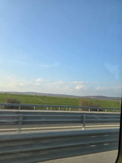 The image displays a daylight roadside scene in Rupkite, Bulgaria, captured from the perspective of a moving vehicle. The lower third of the frame is occupied by a multi-tiered corrugated metal highway guardrail positioned above an asphalt road surface. Beyond the guardrail, a vast expanse of green agricultural field extends into the midground, bordered by a lighter-colored single-rail barrier and sparse bushes. Distant rolling hills are visible on the horizon, beneath a clear blue sky dotted with scattered white cumulus clouds. No individuals or specific actions are present.