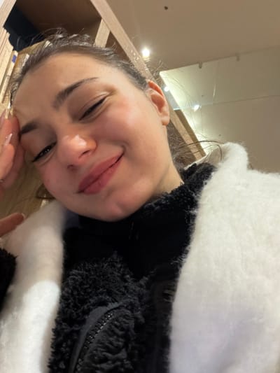 A young woman takes a selfie with a gentle smile. She is wearing a black and white fuzzy jacket. Part of a bookcase is visible in the left side of the frame, along with a ceiling with several lights above. Her nails are manicured, and she appears relaxed.