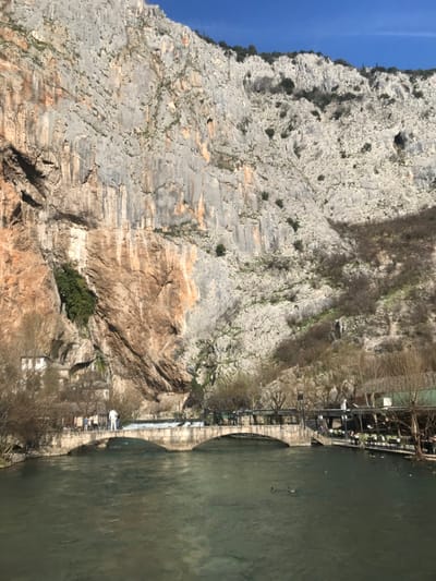 The scene is located in Blagaj, Bosnia and Herzegovina, at the confluence of the Buna River and a dramatic limestone cliff. A massive, textured limestone cliff face, exhibiting grey, tan, and reddish-brown striations, dominates the upper two-thirds of the image under a clear blue sky. Sparse green vegetation grows on ledges and crevices of the cliff.
In the lower third, the turquoise-green Buna River flows from left to right. A three-arched stone bridge spans the river horizontally in the mid-ground. On the bridge, a white human-shaped statue stands prominently near the center, surrounded by approximately seven individuals. To the left of the bridge, partially nestled against the cliff face, are the white-walled structures of the Blagaj Tekke, with a distinct brown rock formation directly above them. A small waterfall or cascade is visible near the Tekke.
To the right of the bridge, along the riverbank, several outdoor dining structures are visible, characterized by green awnings and tables. Multiple individuals are seated or standing in these areas. Bare deciduous trees and shrubs line the riverbanks. Approximately three ducks are swimming on the river's surface. The scene is illuminated by direct sunlight, indicating daytime conditions.