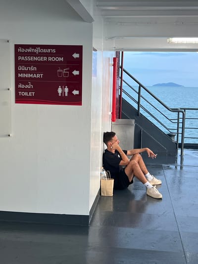 A young Caucasian male with light brown hair and a light beard is sitting on the floor of a ferry, leaning against a white wall. He is wearing a black t-shirt, black shorts, white socks, and beige sneakers. He has a cigarette in his right hand and is looking towards the right. A brown paper bag and a plastic water bottle are on the floor to his left. A sign on the wall to his left indicates directions to "PASSENGER ROOM," "MINIMART," and "TOILET" in both Thai and English. The ferry is moving through the water, with the ocean and a distant island visible. The deck of the ferry is wet, with reflections on the dark grey surface. The lighting suggests it is daytime, possibly late afternoon or early morning, given the soft light and overcast sky. The location is on a ferry, and given the Thai language on the sign, it is likely in Thailand. Specifically, the ferry might be departing from or arriving at Thong Nian, Thailand, or passing through its waters.