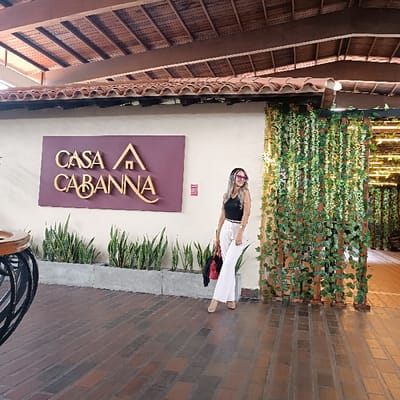 A woman in a black top and white pants stands in front of a building. The building has a sign that says "Casa Cabanna" with a small house icon. A row of green plants grows in a planter below the sign. To the right, an entrance is decorated with hanging vines. The ground is covered with brick. The woman wears sunglasses and carries a red purse. The building's roof is visible at the top of the frame.