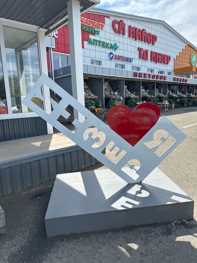 A frontal medium shot captures a large metal sculpture shaped like a diamond, with the Cyrillic letters "ИНЗЕР" cut out and a red heart placed behind the letters "ЗЕ". The sculpture is located in front of a building with the words "СЙ ИНЗЕР" prominently displayed in large red lettering. Below this, signs for "МАРКЕТ", "АПТЕКА+" and "АВТОМАТ" are visible, along with "ПИХТОВОЕ МАСЛО". Stalls are set up under a canopy in front of the building, displaying goods, and people can be seen browsing. The sculpture is situated on a paved surface, with shadows cast by the sun. The overall scene appears to be an outdoor market area in the city of Inzer, Russia.