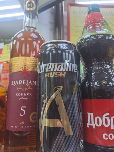 This image presents a close-up view of three distinct beverage containers displayed on a retail shelf in a store located in Меткомелино, Russia. Overhead fluorescent lighting is reflected on the bottles and can, indicating an indoor commercial setting.
From left to right, the objects are:
1. A partial clear glass bottle containing an amber liquid. A blurry background element suggests an "18+" age restriction sign.
2. A sealed 5-year-old bottle of Darejan Cognac, a product of Georgia. This clear glass bottle features a dark maroon label with gold decorative elements, displaying "DAREJAN," "коньяк" (cognac), and the numeral "5" in white text. The bottle is sealed with a dark cap adorned with a gold emblem.
3. A black Adrenaline Rush energy drink can. The can is marked with "АБСОЛЮТНАЯ ЭНЕРГИЯ" (Absolute Energy) at the top and "Adrenaline RUSH" in prominent silver lettering. A large gold "A" symbol with silver diagonal stripes is central to its design.
4. A dark plastic bottle, partially visible on the right side of the frame, sealed with a red screw cap. This bottle contains a dark liquid, consistent with cola, and features a textured surface with small circular indentations. A red label with white Cyrillic text "Добр" and "СО" (indicative of Dobry Cola) is present.
The background is out of focus, showing indistinct retail shelving and a yellow sign with partially visible Cyrillic text. No individuals or active interactions are depicted.