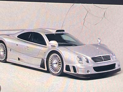 The image is a close-up, angled shot of a silver Mercedes-Benz CLK GTR race car, likely a model or a toy, positioned on a textured, light-colored surface that could be pavement or a studio backdrop. The car is viewed from the front-right side, showcasing its aerodynamic design, including a prominent rear wing, side scoops, and a roof-mounted air intake. The lighting is bright, highlighting the metallic sheen of the car's paint. In the background, a white, textured wall with faint, dark, crack-like lines is visible. The image is of moderate quality, with visible pixelation. There are no people or any other objects in the frame besides the car and the background. The location in Paczkowo, Poland, is not identifiable from the visual information present in the image.