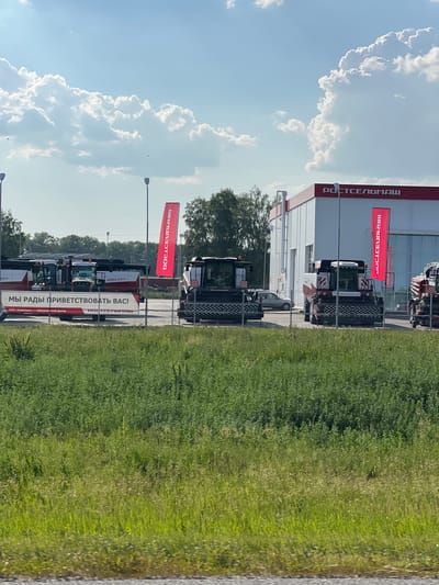 The image depicts an outdoor agricultural machinery dealership on a sunny day with scattered clouds. The foreground is dominated by a lush green grassy field. In the midground, behind a metal fence, several large agricultural machines, specifically harvesters and tractors, are parked. These machines are primarily white and red, with some bearing logos. A prominent red banner with white text in Cyrillic, reading "РОСТСЕЛЬМАШ," is visible on the right side of the image. Adjacent to the machinery is a modern building with a red-trimmed roof, also featuring the "РОСТСЕЛЬМАШ" logo in white letters. A smaller red banner with similar Cyrillic text is attached to a lamppost in front of the building. A car is parked to the left of one of the harvesters. The background consists of trees and a bright blue sky with large, fluffy white clouds. The text on the banner in front of the machines reads "МЫ РАДЫ ПРИВЕТСТВОВАТЬ ВАС!", meaning "WE ARE GLAD TO WELCOME YOU!". The text on the smaller banner reads "РОСТСЕЛЬМАШ" vertically. The scene suggests a commercial display of agricultural equipment. Based on the company name "РОСТСЕЛЬМАШ" and the nature of the equipment, this is likely a dealership located in Russia.