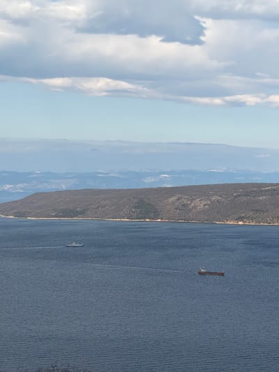 The image presents a body of water bordered by land, potentially a lake or sea near Zagorje, Croatia. In the distance, a mountain range stretches across the horizon under a sky filled with soft, billowy clouds. The water's surface shows slight ripples, suggesting a gentle breeze. Two ships are visible on the water, one longer vessel and a smaller, faster boat leaving a wake. The coastal land features scrub vegetation.