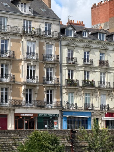 The image shows a multi-story building facade in Nantes, France. The building features a mix of architectural styles, with decorative balconies, dormer windows, and a red brick chimney. The lower levels of the building house several shops, including "EVERYWEAR" and "L'EPT DU FERMIER". A stone wall and lush green trees are visible in the foreground, suggesting the building is situated near a body of water or a park. The sky above is partly cloudy.