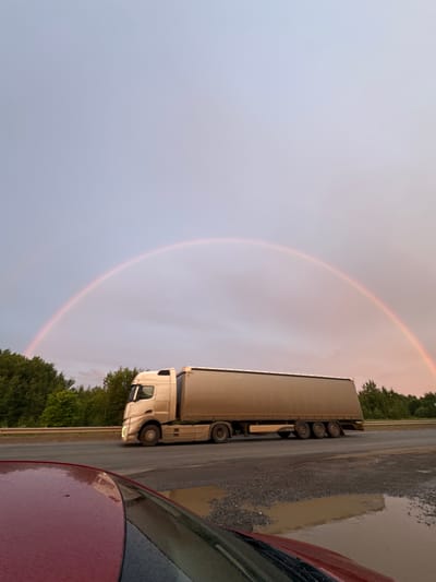The image depicts a truck parked on the side of a road, with a rainbow arching across the sky. The truck is a semi-trailer truck, predominantly white and beige, with multiple wheels visible. The scene appears to be an outdoor setting, possibly a rest stop or a roadside pull-off, given the presence of gravel and a puddle of water on the ground. Lush green trees line the edge of the road in the background. In the foreground, a portion of a red vehicle is visible, suggesting the photo was taken from inside or beside another car.
The sky is overcast and appears to be either early morning or late evening, indicated by the soft light and the presence of the rainbow, which often forms after rain or during misty conditions. The rainbow itself is a prominent feature, a gentle arc of pastel colors against the muted sky. The overall mood is serene and natural, highlighting the beauty of the weather phenomenon. There is no visible text in the image. The location context is Sovkhoznyi, Russia.