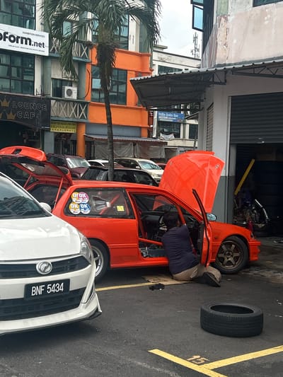 The image captures a daytime scene on a commercial street, likely a workshop or service area, in Subang Jaya City Council, Malaysia. In the foreground, a white car with the license plate "BNF 5434" is parked. Adjacent to it, a bright orange, seemingly modified or race car, is undergoing maintenance. Its hood is fully open, revealing the engine bay, and the driver's side door is also open. A person, presumably a mechanic, is bent over and working inside the vehicle. A detached car tire lies on the asphalt to the right of the orange car, near a yellow road marking numbered "15".
In the background, multi-story commercial buildings line the street, featuring various businesses. A prominent palm tree stands between the cars and the buildings. Visible signs include "EXHIBITION SERVICES," "Form.net," with smaller text "DIKENDALI OLEH MEROFORM ASIA PACIFIC SDN BHD," "LOUNGE CAF," and "Tadika Bucion Eras" (likely a kindergarten). The orange car's rear window displays several circular "OK OK" stickers. The scene suggests an active repair or preparation for the modified vehicle.