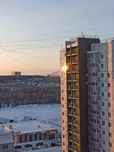 A high-angle view shows a tall building with rows of windows, some reflecting sunlight. Adjacent is a street lined with shops and parked cars, covered with snow. A dense forest stretches beyond, also blanketed in snow. The sky is light, with a gradient of blue and orange, suggesting sunrise or sunset. Power lines are visible across the sky.