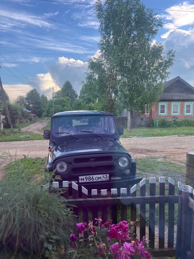 The image depicts a dark green UAZ-469 off-road vehicle parked on a dirt road in a rural area, likely the village of Pigozino, Russia. Two individuals, partially visible through the windshield, are seated inside the vehicle. The car's license plate reads "M986OM 43 RUS". In the foreground, a dark wooden fence with white-capped pickets separates the viewer from the car. Lush green bushes and vibrant pink peonies are visible in front of the fence. Behind the vehicle, a dirt road winds past a traditional Russian wooden house with green trim around its windows. Trees and a vast, cloudy sky are visible in the background, suggesting an outdoor, possibly summer, setting.