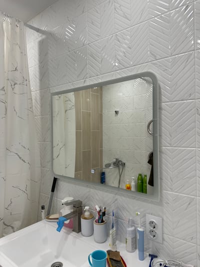 The image shows a modern bathroom interior, likely in a residential setting in Mariinsky, Russia. The walls are tiled with white, textured tiles arranged in a chevron pattern. A large mirror with a rounded frame hangs on the tiled wall above a white sink. The sink area is equipped with a chrome faucet, various toiletries including soap dispensers, toothbrushes in a holder, and toothpaste. On the counter, an electric toothbrush and an electrical outlet with two sockets are visible. A white shower curtain with a marble pattern is partially visible on the left side of the image. The lighting appears to be from overhead fixtures, casting a bright, even light across the scene. The overall impression is one of a clean, functional, and contemporarily designed bathroom.