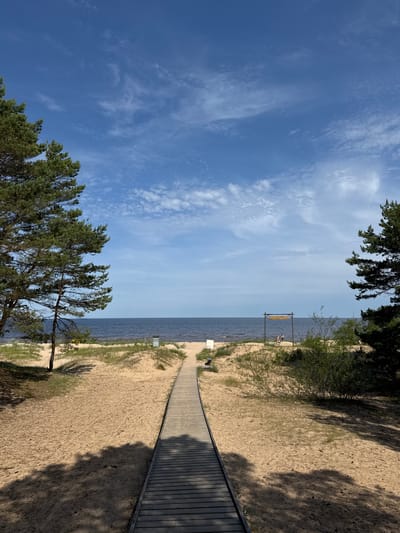 The image depicts a scenic beach on a sunny day in Kalngale, Latvia. The main subjects are the sandy beach, the calm body of water, and the clear blue sky with scattered clouds. A wooden boardwalk curves along the edge of the sand, leading towards the water. On the beach, a few people are visible, appearing to be relaxing.

A notable object is a wooden archway or frame standing near the water's edge. A sign with text is hanging from this structure, but the text is not legible in the image. The presence of trees and dune grass suggests a natural coastal environment. The lighting indicates it is daytime, likely late morning or early afternoon, with the sun casting shadows from the trees onto the sand. The overall mood is peaceful and serene, suggesting a leisurely day at the beach.