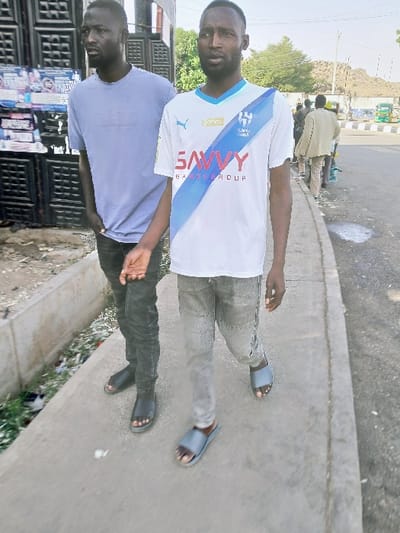 Two young men are standing close together on a paved surface. The man on top wears a white jersey with blue accents, light grey pants, and blue flip-flops. The man on the bottom wears a light purple shirt, dark pants, and black sandals. A wall with posters is behind the second man. A paved road with other people is in the background.