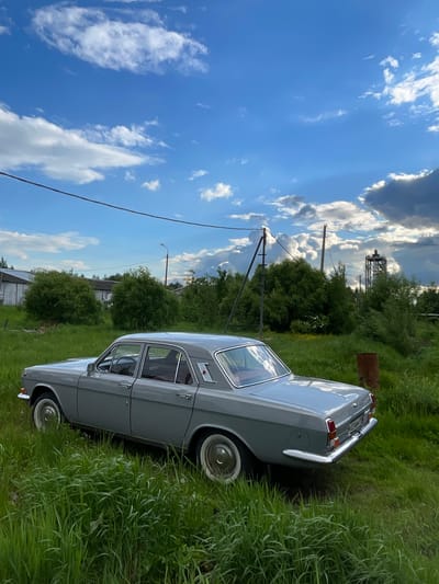 The image depicts a gray vintage car parked in a grassy field in Khar'ino, Russia. A water tower and utility poles are visible in the background against a blue, cloudy sky. A small structure can also be seen further in the background. The car is partially obscured by tall green grass.