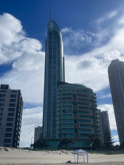 The image captures a beach scene in Surfers Paradise, Australia, dominated by a cluster of modern high-rise buildings against a bright blue sky with scattered clouds. In the foreground, a sandy beach shows footprints and a few items of personal belonging. A portable beach tent with blue and white stripes is set up on the sand, and a person can be seen lying down near it. Behind the beach, several tall, contemporary buildings are visible, including a very tall, slender skyscraper with a curved glass facade and a spire, and a wider, curved building with balconies. Other buildings, varying in height and architectural style, are also present, some appearing under construction or with visible external scaffolding. Trees and other vegetation are sparsely scattered between the buildings and the beach. The overall impression is a modern urban beach environment on a clear day.