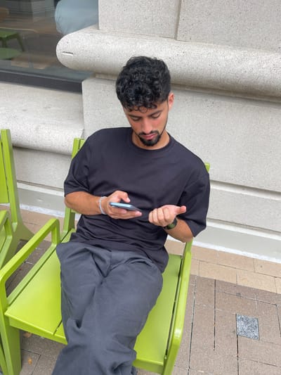 A male individual is seated outdoors on a lime green metal chair, facing forward and looking down at a light blue smartphone held in his left hand. He wears a black short-sleeved t-shirt, dark grey trousers, a thin silver chain, a light blue braided bracelet on his left wrist, and a dark-colored smartwatch on his left wrist. His right hand rests open, palm facing upwards, fingers slightly curled. A second identical lime green chair is partially visible to his left. The background consists of a light grey, textured stone or concrete building facade featuring horizontal banding and a curved architectural element. The ground is paved with rectangular tiles in shades of brown and grey, interspersed with darker grey square tiles. The specific location within Arlington, United States, cannot be determined from the visual information provided.