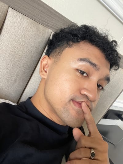 An image captures a headshot of a young adult male in an indoor setting, likely a bedroom or lounge area. He is positioned leaning back against a light beige, textured fabric headboard or cushion. The male has a medium-dark complexion, dark eyes, and dark, tightly curled hair that is shorter on the sides and top, consistent with a fade haircut. Faint stubble is visible on his chin and jawline. He is wearing a black, short-sleeved top with a visible collar line. His right hand is raised, with the index finger placed vertically against his lower lip and upper chin area. A silver-colored ring, featuring a dark, possibly oval-shaped, stone or inlay surrounded by ornate metalwork, is worn on the ring finger of his right hand. His gaze is directed forward, slightly upwards, towards the camera. The background behind him includes the upholstered headboard, a lighter-colored wall above it, and a dark wooden or laminate surface to the upper right. The image provides no specific visual cues to confirm its geographic location within Kayuambon, Indonesia, or any other specific region.