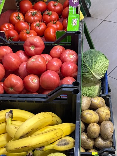 The image displays a produce section within a market or grocery store located in Suhache, Bulgaria. The foreground and midground are filled with various fresh fruits and vegetables organized in stacked plastic crates.
In the upper two-thirds of the frame, several black plastic crates are densely packed with red tomatoes, many exhibiting visible green stems. A partial cardboard carton, green and purple, with "SOY" branding, is positioned among the tomatoes in the upper-middle section.
To the right, a dark blue or black crate contains two heads of green cabbage, the upper one larger and more prominent with distinct leaf texture. Below this, another dark crate is filled with numerous light brown potatoes, some with visible dirt remnants and yellowed areas on their skins. A small yellow price tag is partially visible within the potatoes.
In the lower left, a black plastic crate holds a quantity of ripe yellow bananas, some displaying minor brown spotting. The environment includes a light gray tiled floor with dark grout lines, visible along the right side of the image. No individuals are present, and no actions or interactions are depicted.