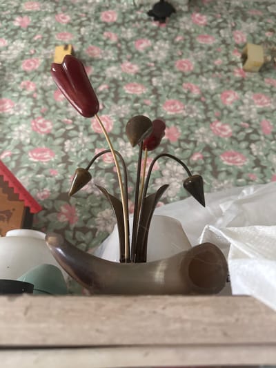 The image depicts an arrangement of decorative metal flowers, with red and brown accents, in a horn-shaped vase. The arrangement is placed on a surface with a floral pattern. Other objects are visible around the vase, partially obscured by a white cloth or bag. The setting appears to be indoors. The location is Студзёнкаўскі сельскі Савет, Belarus.