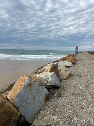 The image depicts a coastal scene with a rocky breakwater and a paved walkway. The breakwater is composed of large, irregular rocks, some of which are tinged with orange and grey. Beyond the breakwater, the ocean stretches to the horizon under a cloudy sky. Gentle waves are breaking on a sandy beach to the left of the breakwater. The walkway, made of gravel, runs along the right side of the breakwater, leading into the distance. A signpost with multiple warning symbols is visible on the walkway, near the breakwater. A lone figure is walking on the walkway in the far distance. This scene is consistent with the Muttonbird Island Nature Reserve in Coffs Harbour, Australia, known for its breakwater walkway and ocean views.