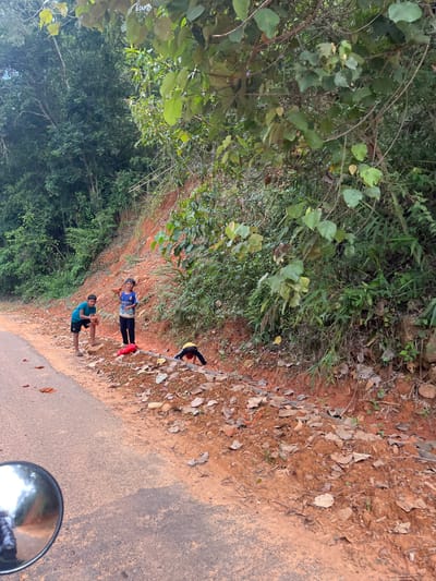 This is a medium shot, eye-level, outdoor photograph taken on a sunny day in Son Dien, Vietnam. The image captures a roadside scene with three children and a motorcycle mirror in the foreground.
On the left side of the paved road, a young boy wearing a blue t-shirt and black shorts is squatting down. To his right, a slightly older boy in a blue and white jersey and dark pants stands looking towards the camera. Further to the right, a third child, wearing a yellow shirt and red head covering, is partially visible, hunched over near the edge of the road, possibly interacting with something on the ground.
The roadside is composed of red dirt, scattered with dry leaves and some broken pieces of brick. Dense green foliage and trees line the embankment behind the children, creating a natural backdrop. A portion of a motorcycle's circular rearview mirror is visible in the bottom left corner of the frame, reflecting a blurred image of what appears to be the photographer. The overall scene suggests a rural or semi-rural setting.