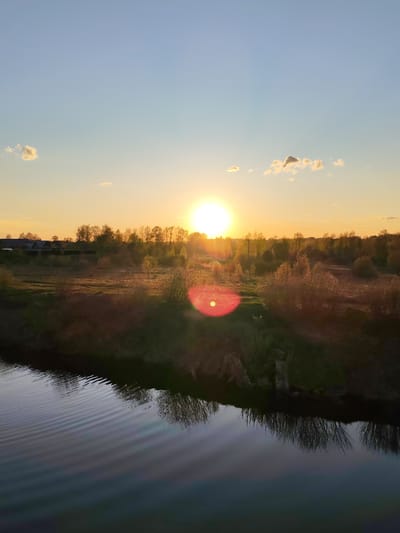 The image is a landscape photograph taken in Myshkin, Russia at sunset. The sun is setting on the horizon, producing a bright orange light on the landscape, with a lens flare visible slightly below. A dark river runs in the foreground, reflecting the landscape above. The land is flat, with trees and bushes in the middle-ground. Buildings are visible in the background. The sky is blue, with a few scattered clouds.