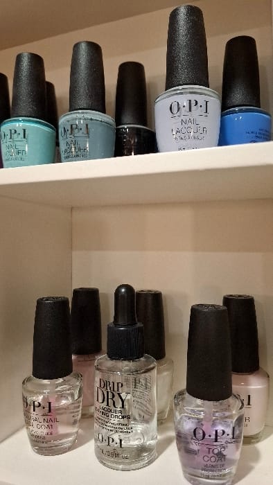 The image displays an indoor scene featuring two white shelves laden with various OPI brand nail care products. The specific location in the city of Podbadac, Montenegro, is not discernible from the visual evidence provided.
On the upper shelf, five OPI Nail Lacquer bottles are arranged. From left to right, these include a teal-colored bottle, a grey-blue bottle, a dark-colored (possibly black or deep brown) bottle, a light lavender bottle, and a bright blue bottle. Each bottle is topped with a textured black cap.
The lower shelf presents a collection of five OPI nail care items. From left to right: a clear bottle containing a pink liquid, labeled "NATURAL NAIL BASE COAT"; a clear bottle with a black dropper cap, containing a clear liquid labeled "DRIP DRY LACQUER DRYING DROPS"; a light-colored (possibly white or very pale) bottle; a clear bottle containing a light purple liquid, labeled "TOP COAT"; and a light pink bottle. The non-dropper bottles on this shelf also feature textured black caps.
No individuals or direct actions are depicted; the objects are static on the shelves. The background consists of the interior white surfaces of the shelving unit.