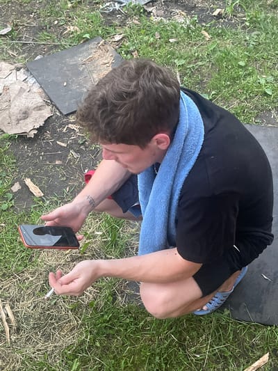 A man is seen from a high-angle perspective, crouching outdoors on grass and dirt. He is wearing a black t-shirt, black shorts, and light blue slides. A blue towel is draped around his neck. He is holding an orange smartphone in his left hand and a lit cigarette in his right hand. The background shows dry leaves, grass, and a dark, flat object. The image is an outdoor scene with no specific urban landmarks visible, making it impossible to determine if it is located in Вымпел Снт, Russia.