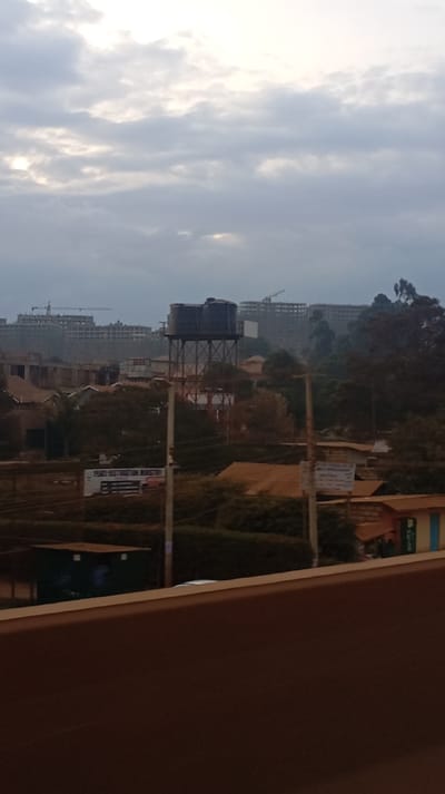 The image depicts a cityscape under a cloudy sky. A large water tank stands prominently amidst the buildings and trees. Construction cranes are visible, suggesting ongoing development. Below, a sign reads "Peace Restoration Ministries." The foreground is slightly obscured, offering a partial view from an elevated position, possibly a balcony.