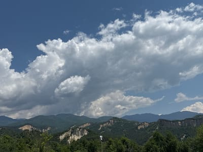 The image displays a vast mountainous landscape under a variable sky near Rozhen, Bulgaria. The upper two-thirds of the frame are dominated by a blend of bright white cumulus clouds and darker grey, heavier clouds, with intermittent patches of deep blue sky visible. A prominent range of dark green, densely forested mountains forms the backdrop, stretching across the horizon. In the mid-ground, significant geological formations, characterized by light-colored, eroded earth pyramids and steep, sandy cliffs, are visible, contrasting sharply with the surrounding dark green forests. These formations appear on both the left and right sides of the frame, interspersed with lush tree cover. The foreground is filled with the dense canopy of dark green trees. In the lower center, a dark pole with metallic attachments, resembling surveillance or communication equipment, rises above the tree line. No human figures or animals are present. The scene is illuminated by natural daylight, with shadows cast by the cloud formations adding depth.