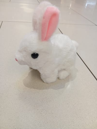 A close-up, high-angle shot features a white, plush toy rabbit with pink inner ears and black eyes. The rabbit is positioned on a light-colored, textured surface that appears to be a tiled floor. The surface has subtle speckling and grout lines are visible. The lighting suggests it is daytime, likely indoors, possibly in a home or retail environment. The scene is static, with no activity taking place other than the presentation of the toy. The focus is on the toy rabbit, with the background showing some blurred floor tiles and a faint shadow of the rabbit. No people, text, or specific weather conditions are discernible. The context indicates the location is Wołomin, Poland, but this is not visually evident from the image itself.