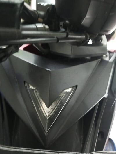 The image focuses on the front of a dark-colored motorcycle. The primary feature is a sharply angled, V-shaped light element embedded in a matte gray, angular fairing. Above, motorcycle handlebars and components are visible, positioned near a gray panel. The overall impression is of a modern, aggressively styled motorcycle front.