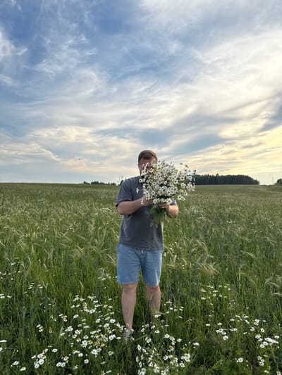 A man stands in a field of green grass and daisies, holding a large bouquet of daisies. He is wearing a grey t-shirt and blue denim shorts. The sky is partly cloudy with wispy white clouds. In the background, a line of trees can be seen on the horizon. The location is identified as Snovitsy, Russia.
