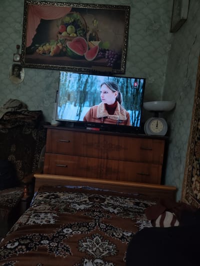 This image depicts an interior scene in a modestly furnished room, possibly located in Yekaterinovka, Russia. The room is cluttered with various items, suggesting a lived-in space. Dominating the scene is a large, framed still-life painting of fruit hanging on a wall adorned with patterned wallpaper. Below the painting, a television screen displays a woman in winter attire.
In front of the television is a wooden dresser with three drawers, each equipped with a silver handle. On top of the dresser, to the right of the television, sits a mechanical scale with a white bowl and a circular dial. To the left of the dresser, a portion of a bed is visible, covered with a patterned blanket. Another piece of furniture, possibly a chair or the armrest of a sofa, is partially visible on the left, draped with blankets and fabric.
The overall impression is one of a slightly dated and somewhat cluttered living space, typical of some homes in smaller Russian towns or villages. The lighting suggests it is daytime, but the room appears to be dimly lit, possibly due to the placement of windows or the time of day.