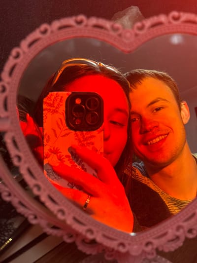 The image depicts a mirror selfie taken indoors in Prikubanskiy, Russia. The reflection is framed by a heart-shaped mirror with a white, intricately decorative border. The scene is illuminated by a strong red-orange light source.
Two individuals are reflected in the mirror. On the left, a female subject with dark hair, wearing a light-colored headband, holds a silver smartphone equipped with a white case featuring a floral pattern and prominent triple camera lenses. The phone covers the lower half of her face. A gold-toned ring with a clear stone is visible on her left ring finger. To her right, a male subject with short, light brown hair and light facial stubble is smiling, revealing his upper teeth. He is wearing a striped undergarment beneath a darker outer garment. In the upper background of the reflection, a faint, light-colored, possibly crystalline object is discernible above the female's head.
