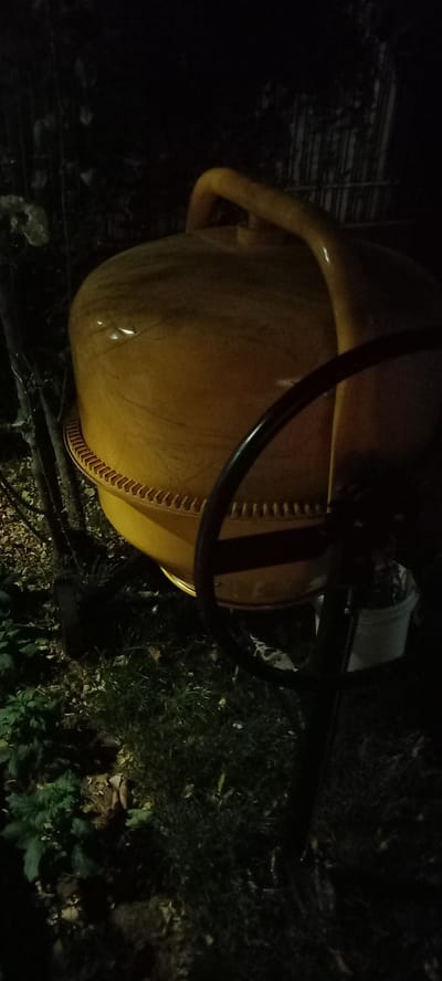 The image depicts a yellow cement mixer positioned outdoors at night in Iskra, Bulgaria. The mixer, a substantial piece of equipment, has a large drum with a visible rim of teeth around its opening, and a curved handle on top. A black steering wheel is attached to the front, suggesting it's a portable model. The mixer is situated on a patch of ground covered with grass and fallen leaves. In the background, indistinct foliage and a fence-like structure are visible, all shrouded in darkness. The lighting appears to be artificial, casting a yellowish glow on the mixer and highlighting some of the surrounding vegetation. A white bucket is partially visible beneath the mixer's drum. No individuals are present in the frame, and no specific actions or interactions are taking place. The environment suggests an outdoor storage area or a worksite after operations have ceased for the day.